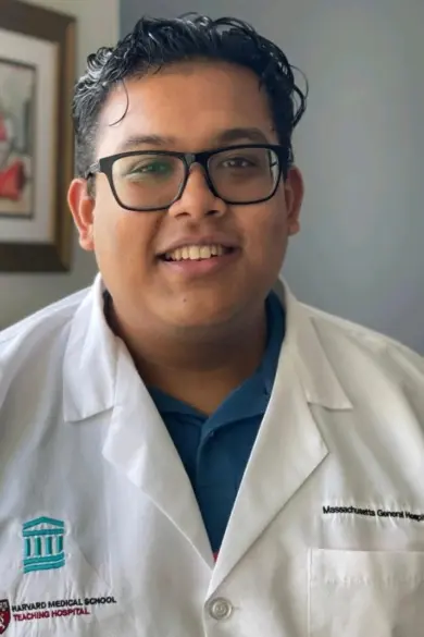 A person wearing glasses and a white medical lab coat stands indoors. The coat has logos from Harvard Medical School and Massachusetts General Hospital. Artworks are visible on the wall behind them.