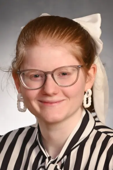 A person with long red hair tied back with a white ribbon and wearing glasses is smiling. They are dressed in a black and white striped shirt and white geometric earrings against a neutral background.
