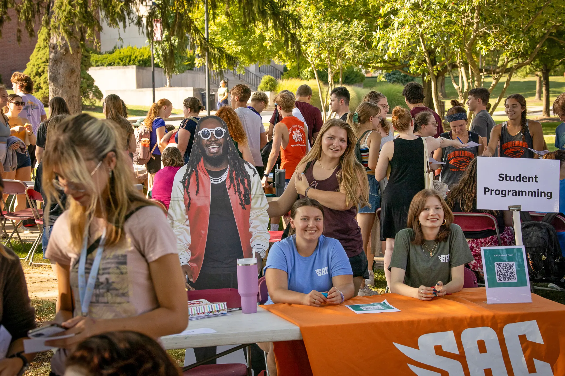 Outdoor college fair buzzing with students at activity booths. Embracing campus life, three smiling people at a "Student Programming" table stand beside a cardboard cutout of a man in sunglasses. Lush greenery envelops the lively event.