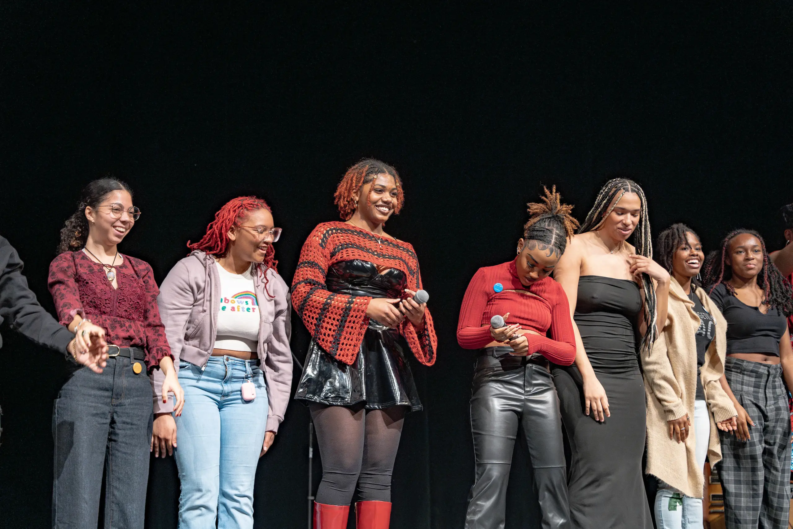 A group of people stands on stage smiling and holding microphones, showcasing their colorful, stylish outfits against a black background. It's a perfect example of "Learn By Doing," as they confidently embrace the spotlight with hands-on experience.
