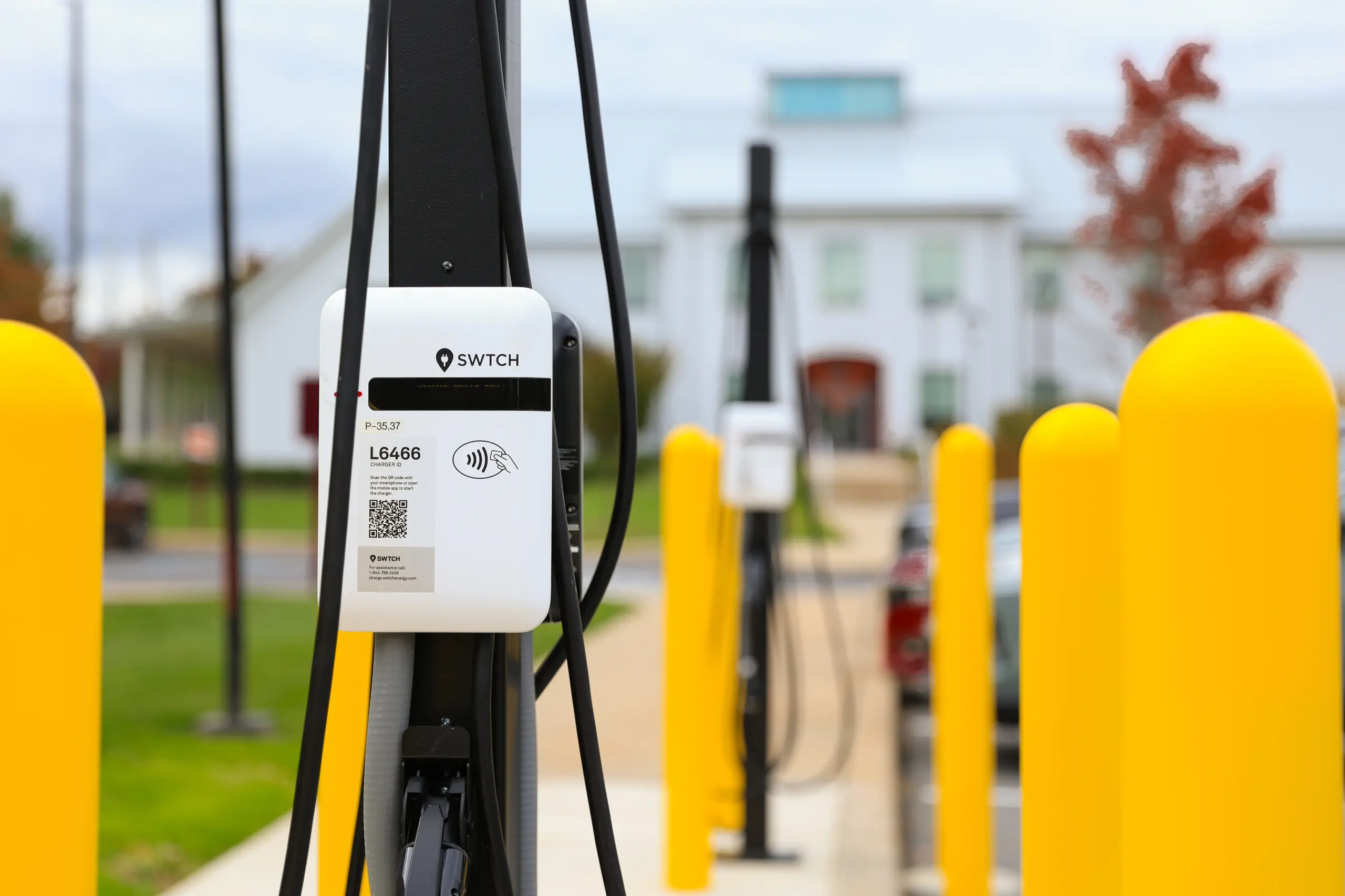 Close-up of an electric vehicle charging station with yellow protective posts, located outdoors on campus with a blurred building and trees in the background, reflecting a commitment to sustainability and modern Campus Life.
