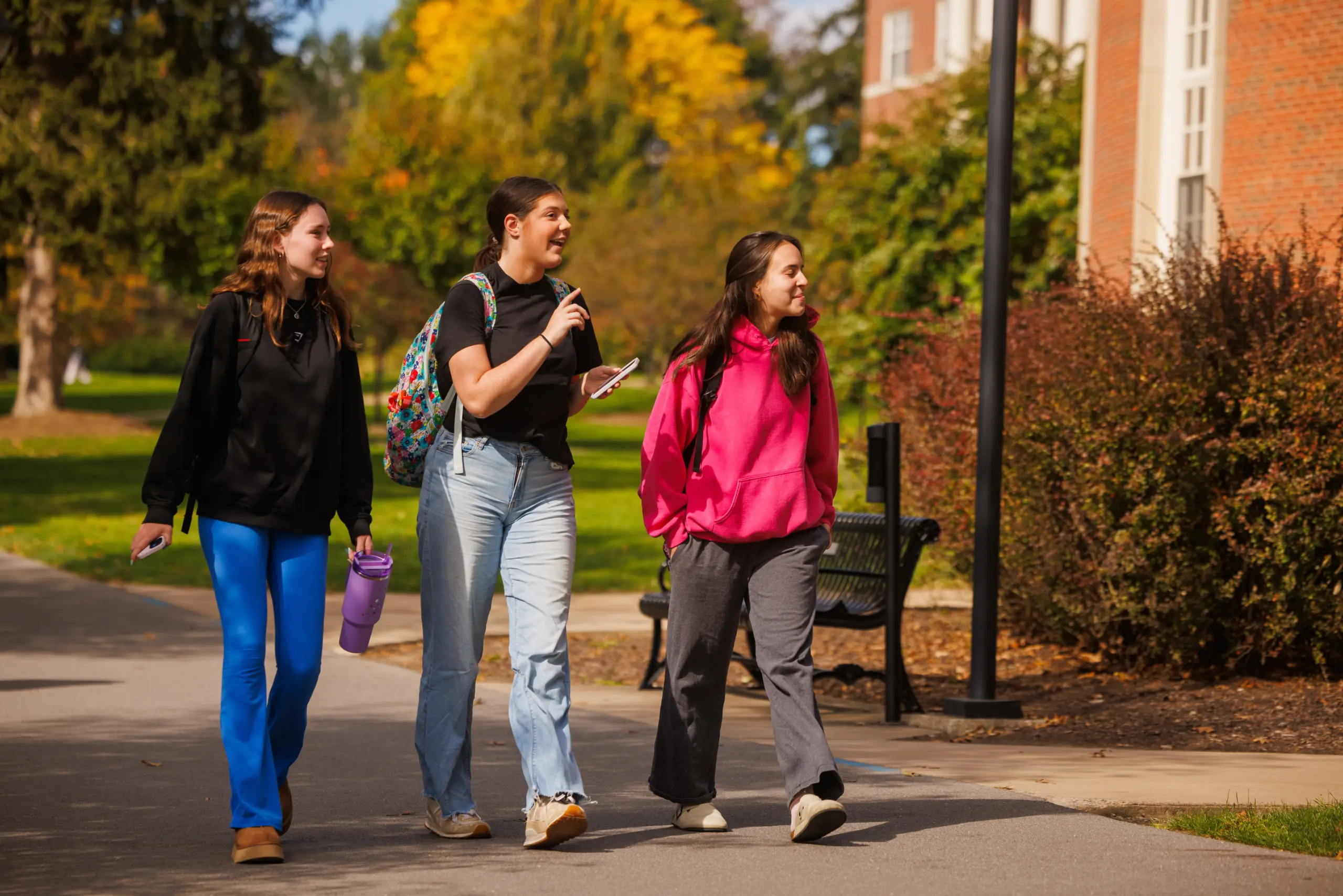 Three young people walk on a campus path in bright sunlight, surrounded by autumn foliage. They are chatting and smiling, wearing casual clothes and backpacks, with one holding a phone and another a water bottle. A brick building is in the background.