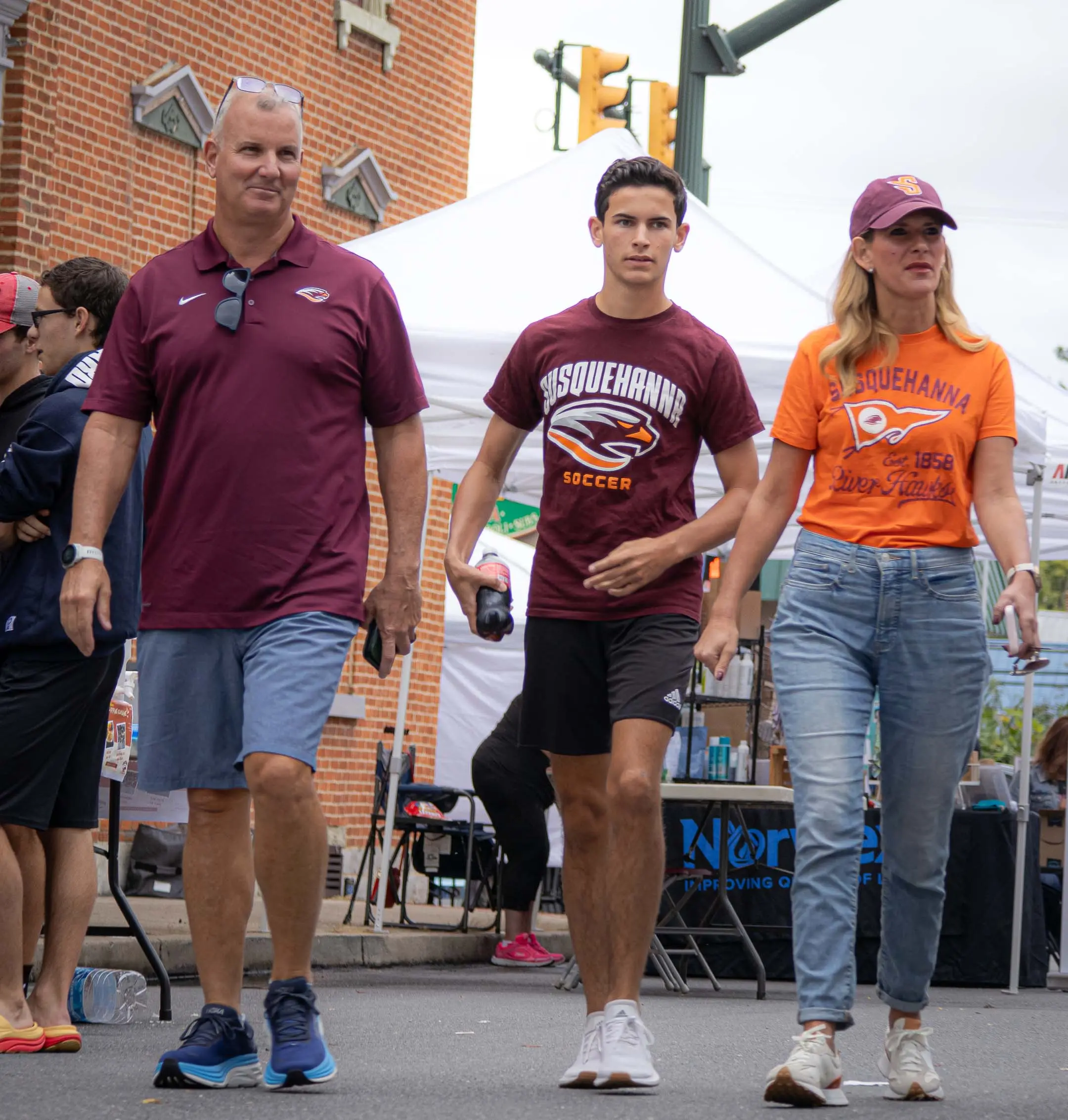 Three people walk together at an outdoor event, all wearing Susquehanna University shirts. They appear relaxed, enjoying campus life with tents and other people visible in the background.