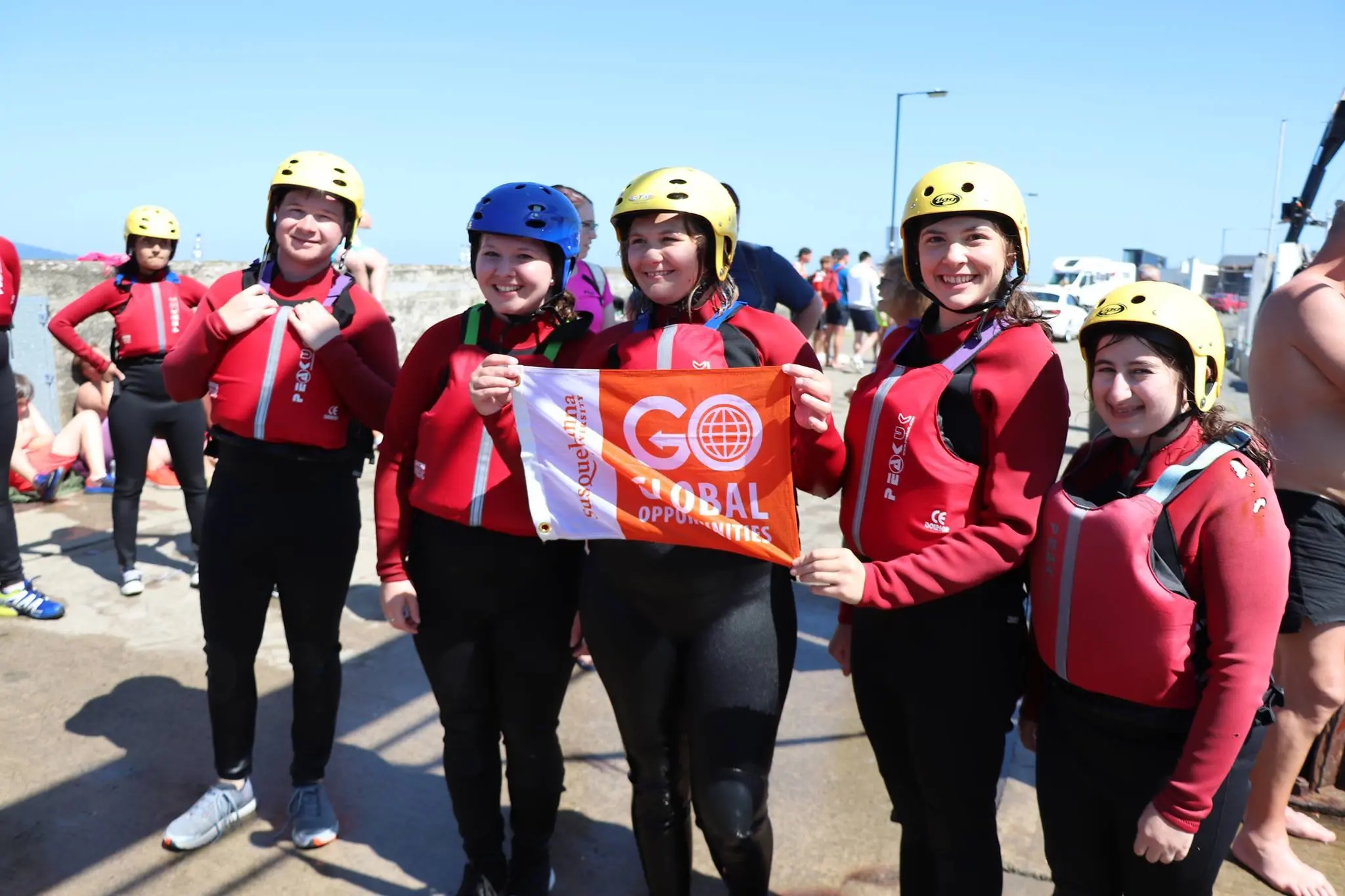 Five people in red wetsuits and helmets stand together outdoors, smiling and holding a "GO GLOBAL OPPORTUNITIES" flag. It's a sunny day with others and equipment in the background—celebrating their GO Study Abroad adventure.
