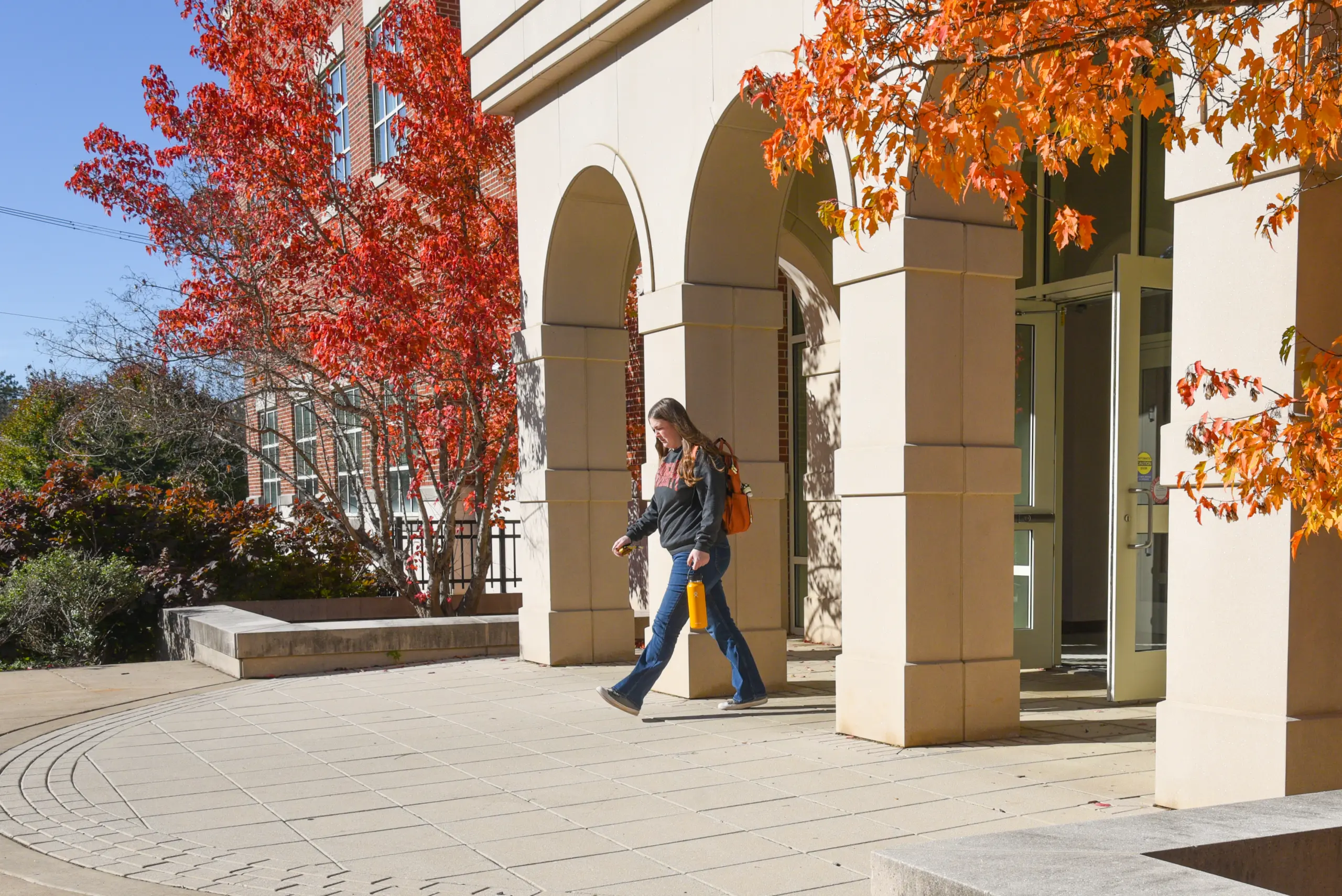 A person walks outside a building with arched doorways, surrounded by vibrant orange autumn trees and sunlight, carrying a bag and a drink bottle.