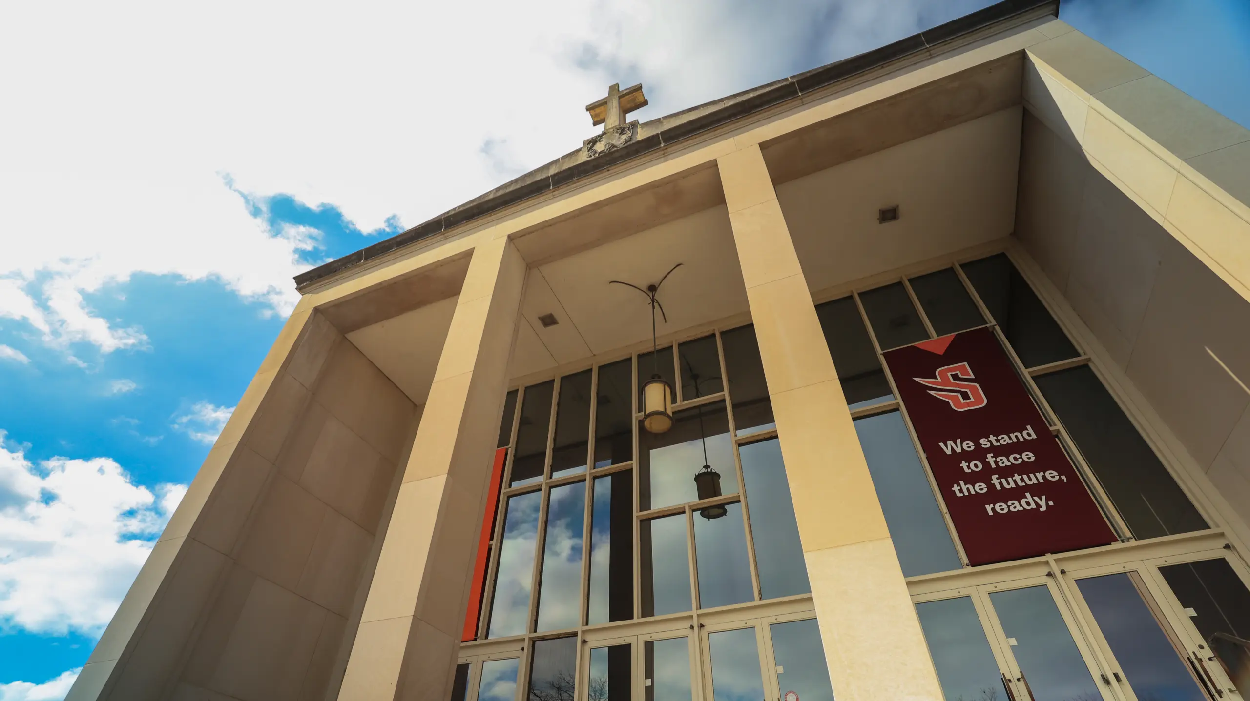 Low-angle view of a building with tall columns, a cross on the roof, large glass windows, and a red banner reading, "We stand to face the future, ready." The sky is partly cloudy.