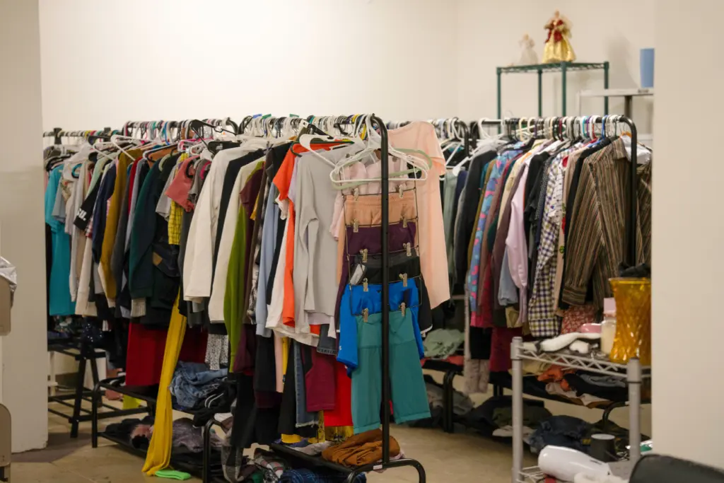 A cluttered clothes rack brimming with colorful shirts, pants, and jackets hints at sustainability initiatives. The dim lighting reveals some folded clothes on the floor and a shelf with additional storage items made from eco-friendly materials in the background.
