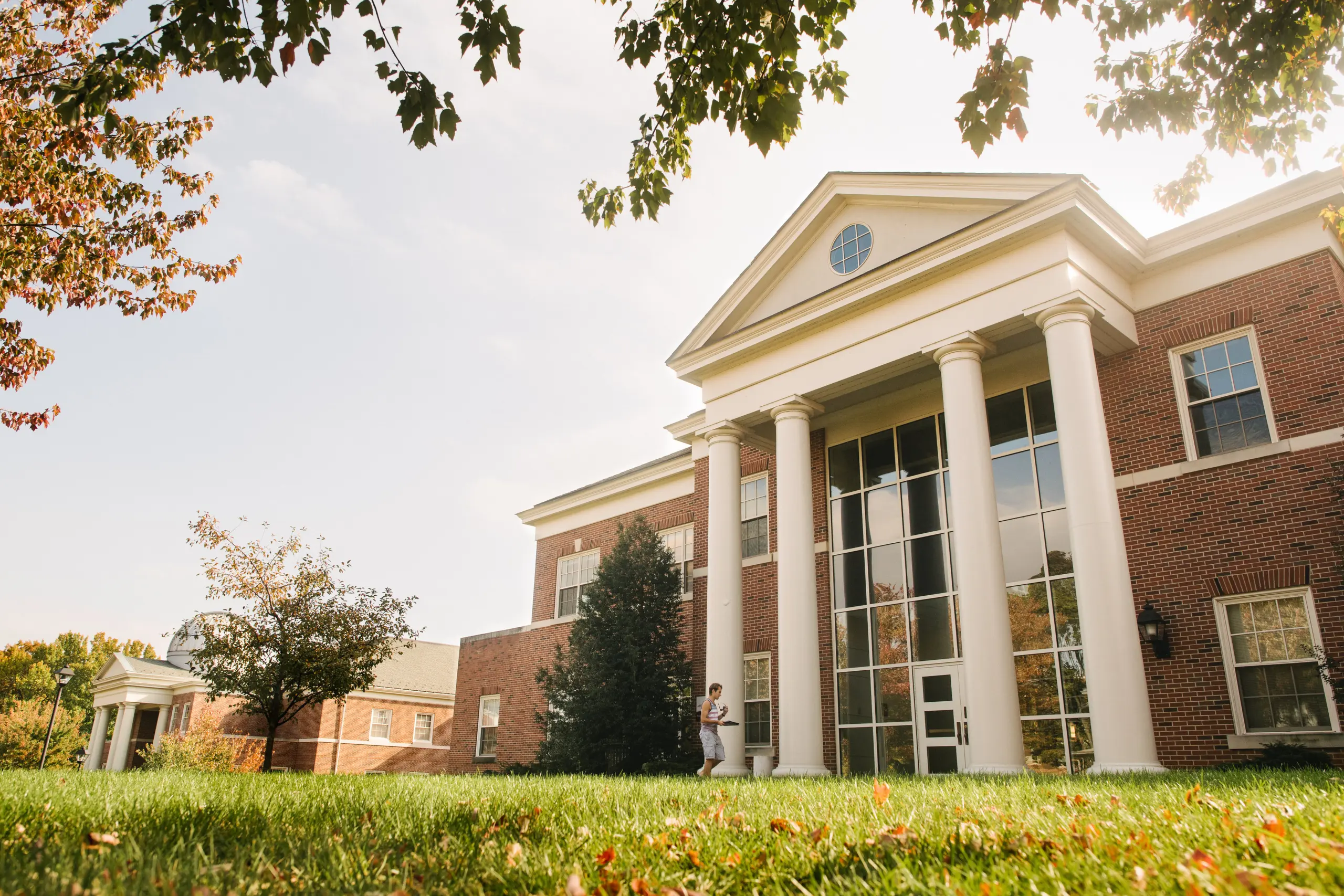 A red brick building with six white columns and large windows sits on a grassy lawn under a partly cloudy sky. Tall trees with green and autumn-colored leaves frame the scene.