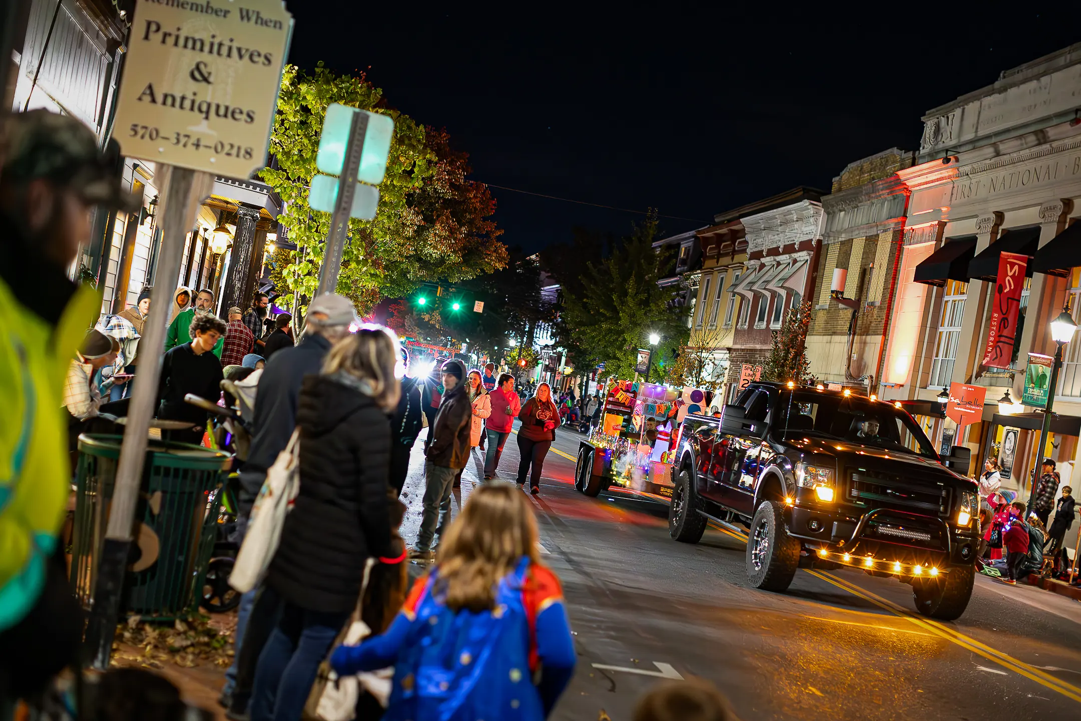 A lively nighttime street parade captures the spirit of campus life, with people in costumes, a decorated pickup truck adorned with lights, and crowds watching from sidewalks while illuminated shops and buildings line the background.