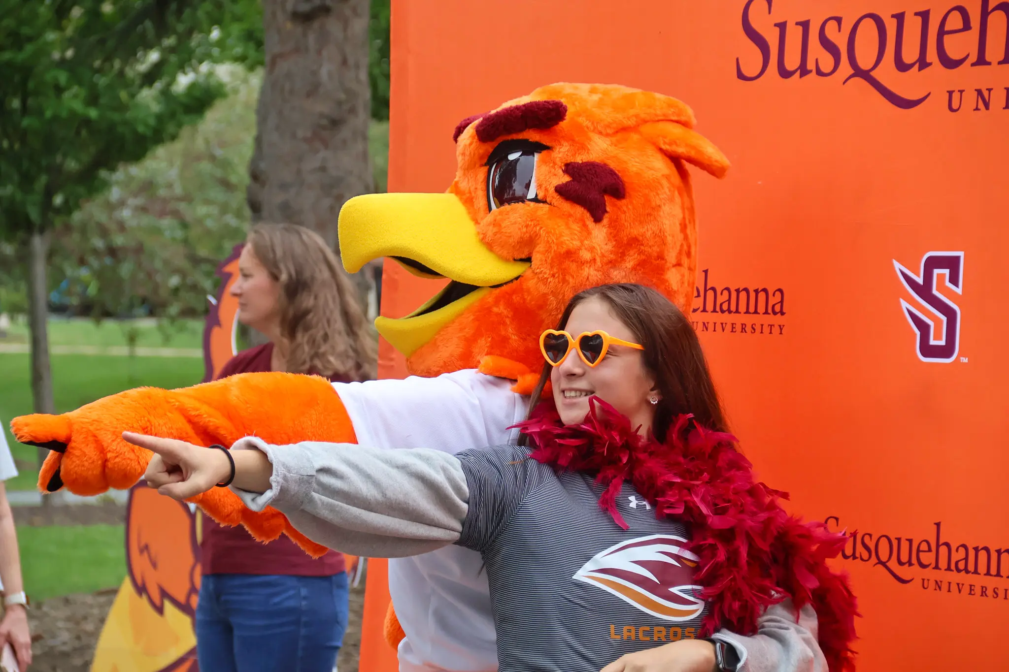 About: A person wearing heart-shaped sunglasses and a feather boa poses with an orange bird mascot; both are smiling, pointing, and having fun in front of a Susquehanna University backdrop.