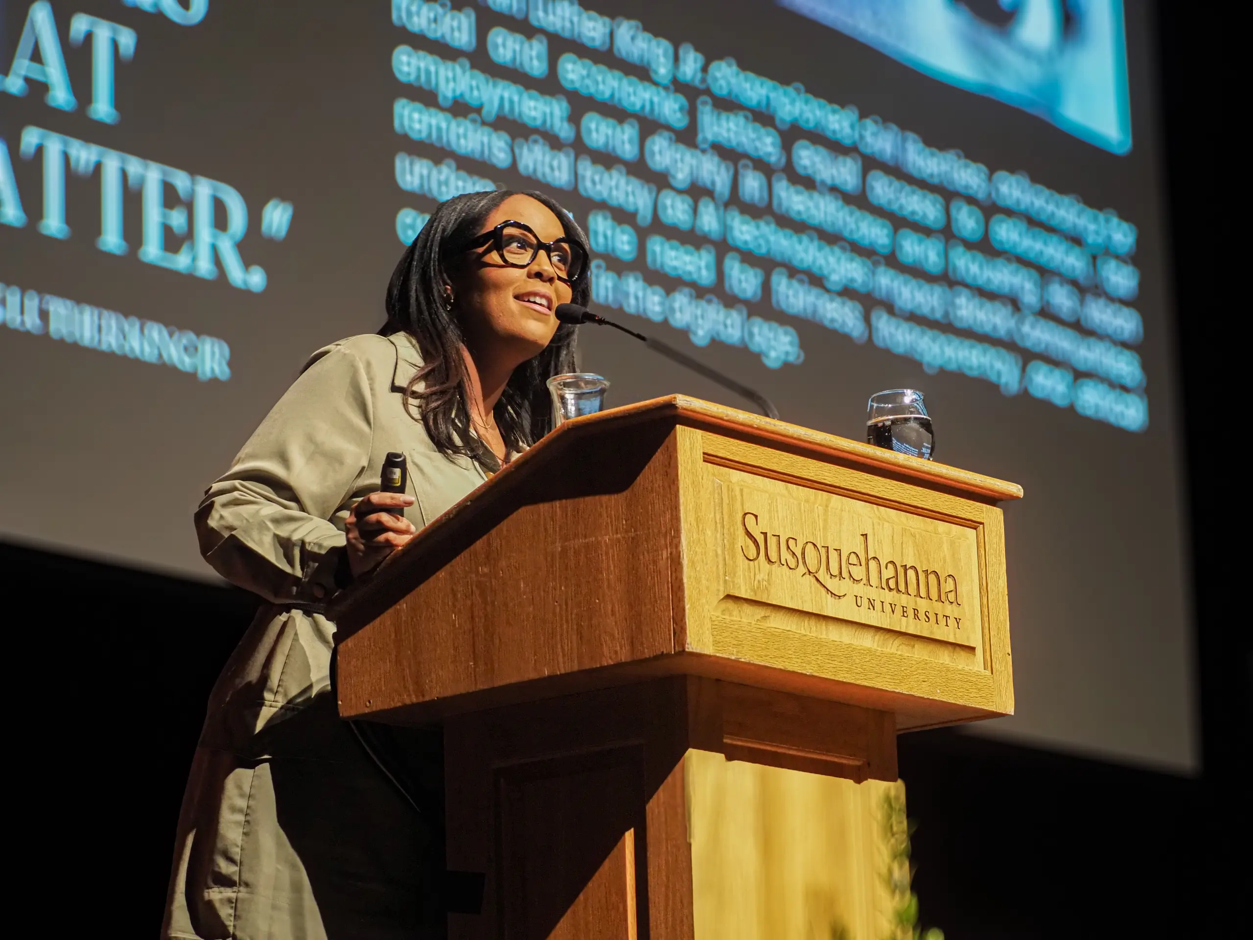 A woman speaks at a wooden podium inscribed with "Susquehanna University" during a presentation about her topic. She wears glasses and stands in front of a screen displaying text and an image.