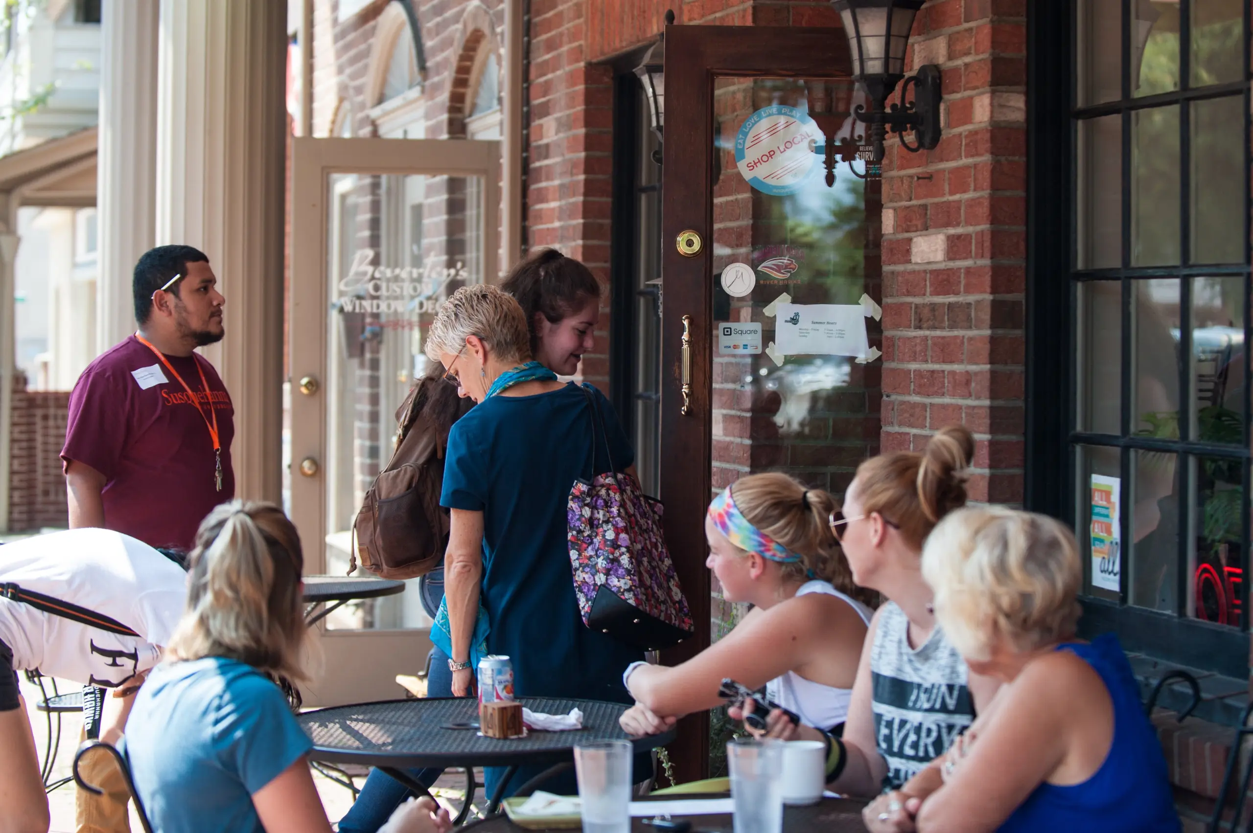 A group of people sit and chat at outdoor tables of a brick-walled café, enjoying campus life. Others stand near the entrance as sunlight brightens the scene and empty glasses are visible on the tables.