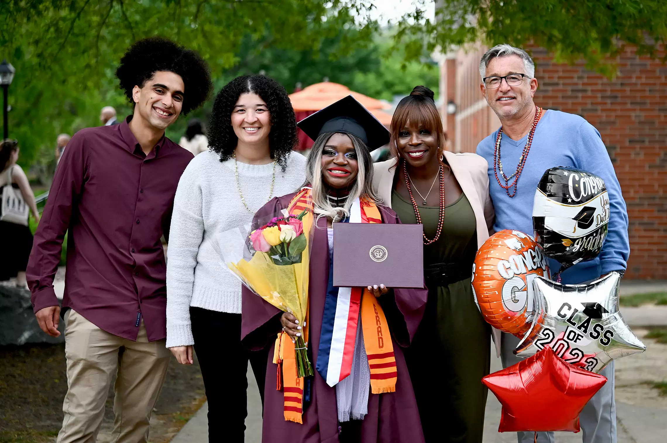 A smiling graduate in cap and gown holds flowers and a diploma at commencement, surrounded by four happy loved ones. They stand outdoors with "Congrats Grad" and "Class of 2023" balloons.