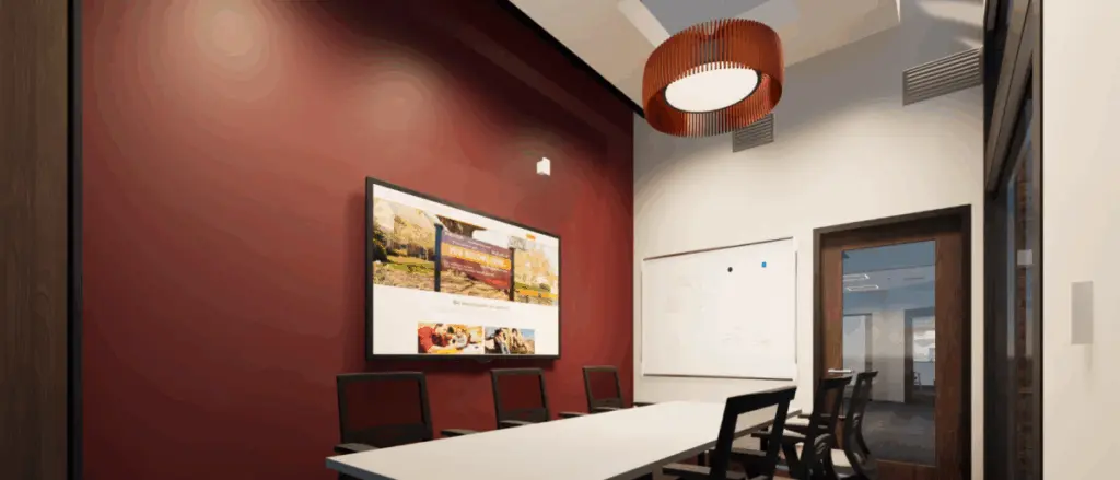 A modern conference room in Building and Spaces featuring a long white table, black chairs, a large wall-mounted screen displaying images, a whiteboard, maroon accent wall, and a large circular ceiling light.
