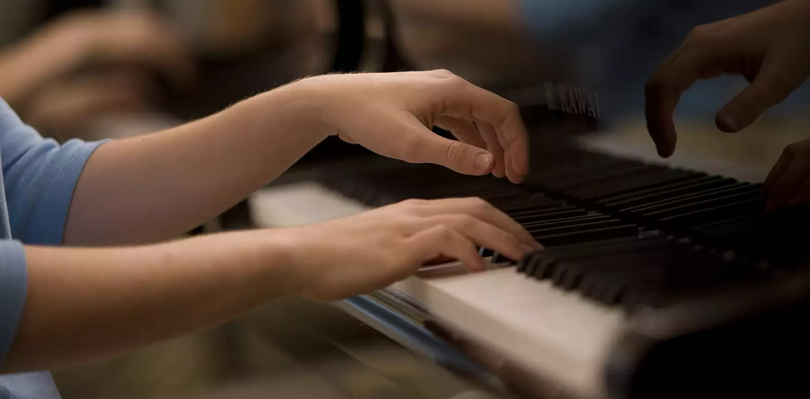 Close-up of a person’s hands playing a black piano keyboard, with reflections of their hands visible on the glossy surface of the piano.
