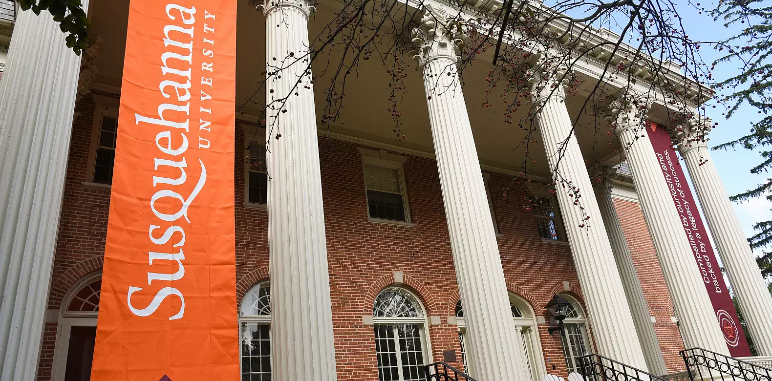 A brick building with tall white columns displays large banners reading "Susquehanna University" and "We will give you the tools to shine." Leafy branches frame the top of the image.