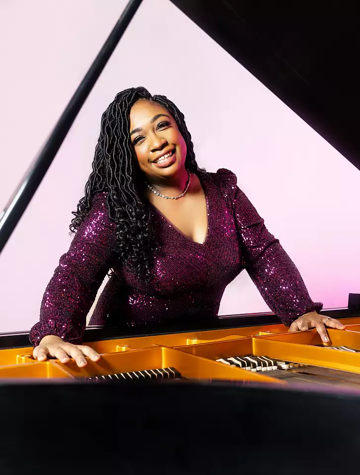 A woman in a sparkly purple dress smiles while leaning over the inside of a grand piano, with its lid open, against a light pink background.