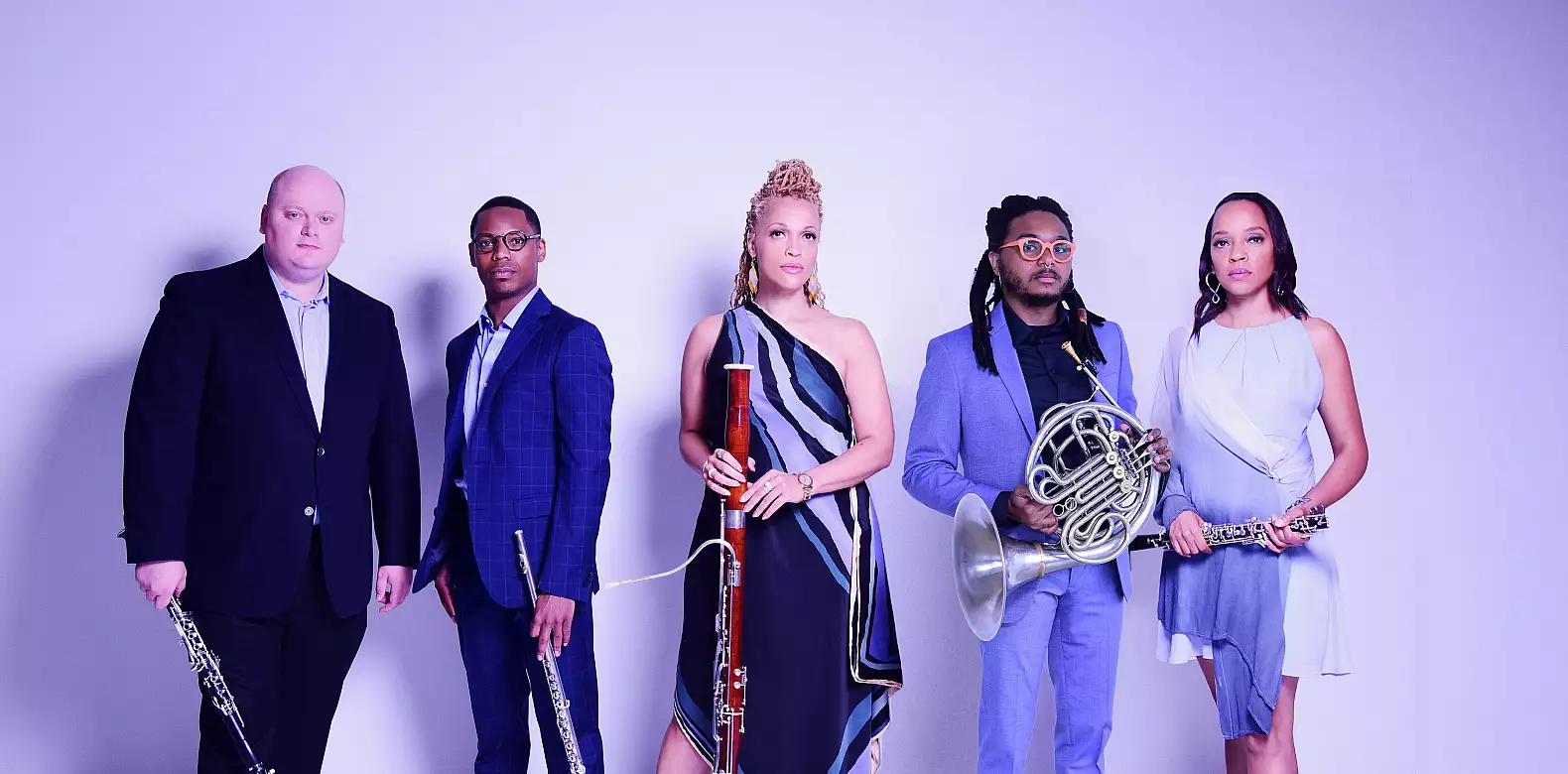 Five musicians stand together holding instruments, including an oboe, clarinet, bassoon, French horn, and flute. They are dressed in formal and stylish attire, posed against a light purple background.