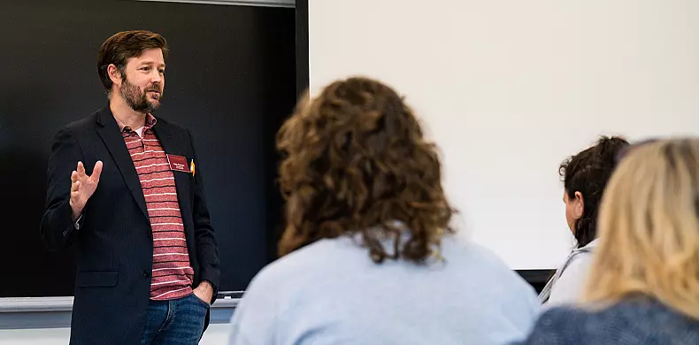 A man in a blazer and striped shirt stands in front of a classroom, gesturing as he speaks to several seated students facing him. A blackboard is visible behind him.