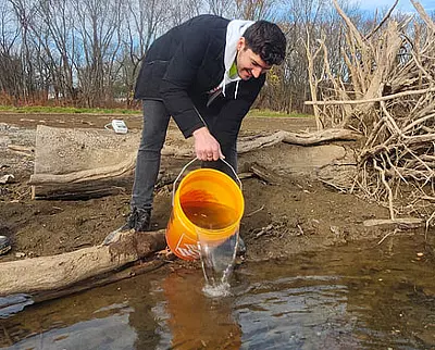 A person in a black jacket pours water from an orange bucket into a stream on a muddy riverbank with fallen logs and leafless trees in the background.