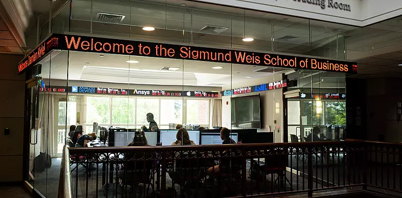 A modern classroom with glass walls shows students working at computers. An LED ticker displays “Welcome to the Sigmund Weis School of Business” above stock and financial information.