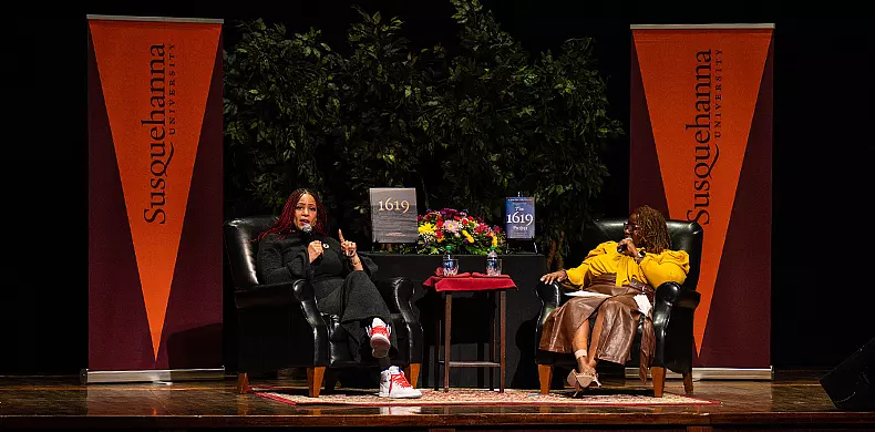 Two women sit on stage in armchairs between two orange Susquehanna University banners, speaking at an event. A table with flowers and books is between them and green plants are in the background.