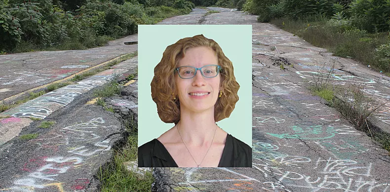 A smiling woman with curly, shoulder-length hair and glasses is shown in a portrait overlaid on a cracked, graffiti-covered road surrounded by overgrown greenery.