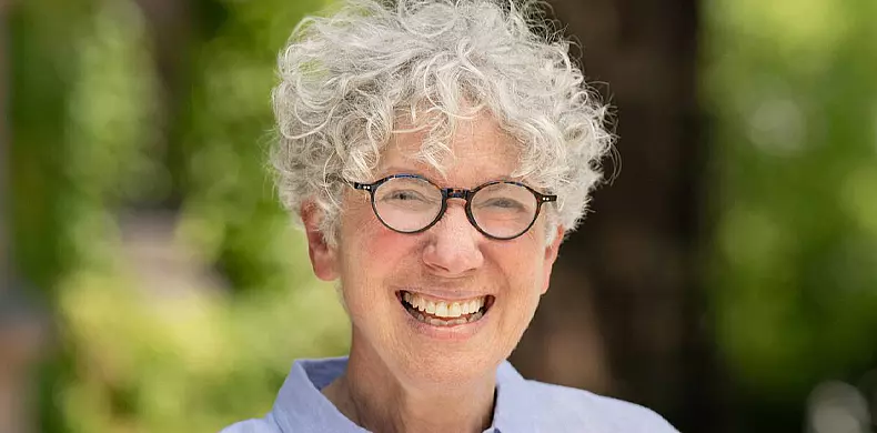 An older person with short, curly gray hair and round glasses smiles warmly outdoors, wearing a light blue shirt. Greenery is blurred in the background.
