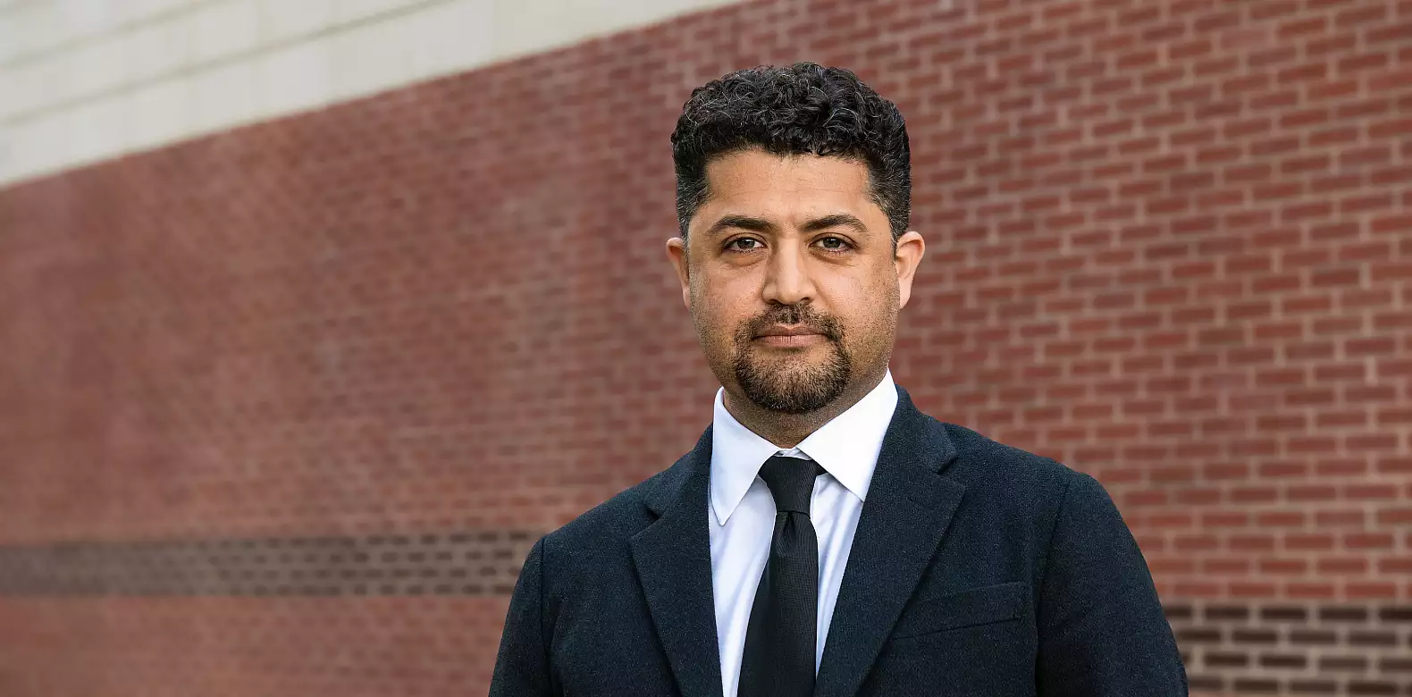 A man with short curly hair and a trimmed beard wears a dark suit, white shirt, and black tie, standing in front of a red brick wall.