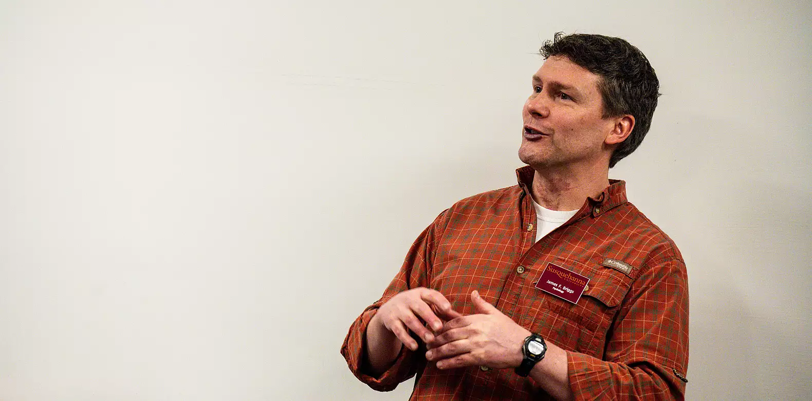 A man wearing a red plaid shirt and a name tag gestures with his hands while speaking, standing in front of a plain white wall.