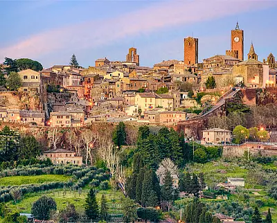 A picturesque hilltop town with historic stone buildings, towers, and a clock tower, surrounded by lush green trees and countryside under a pastel sky at sunset.