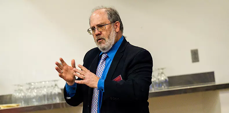 A middle-aged man with glasses, a beard, and thinning hair, wearing a dark suit, blue shirt, and patterned tie, gestures with his hands while speaking in an indoor setting.