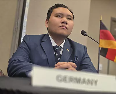 A person in a suit sits at a table with a microphone, a small German flag, and a nameplate labeled "GERMANY." The setting appears formal, possibly a conference or meeting.