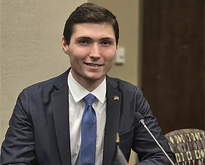 A young man in a dark suit, white shirt, and blue tie sits at a table with a microphone in front of him, smiling at the camera. The background shows a beige wall and a door.