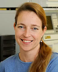 A woman with long light brown hair tied back, wearing a blue top, smiles at the camera. She is indoors, with office or lab equipment visible in the background.