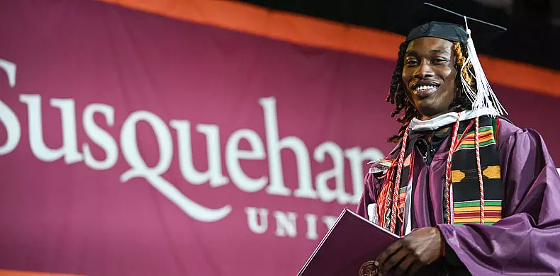 A smiling graduate in a cap and gown holds a diploma in front of a maroon Susquehanna University banner.