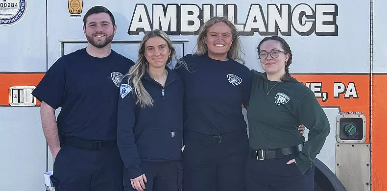 Four smiling people in uniform stand arm-in-arm in front of an ambulance. Three wear navy shirts, one wears a green shirt. The ambulance has "AMBULANCE" written in large letters above them.