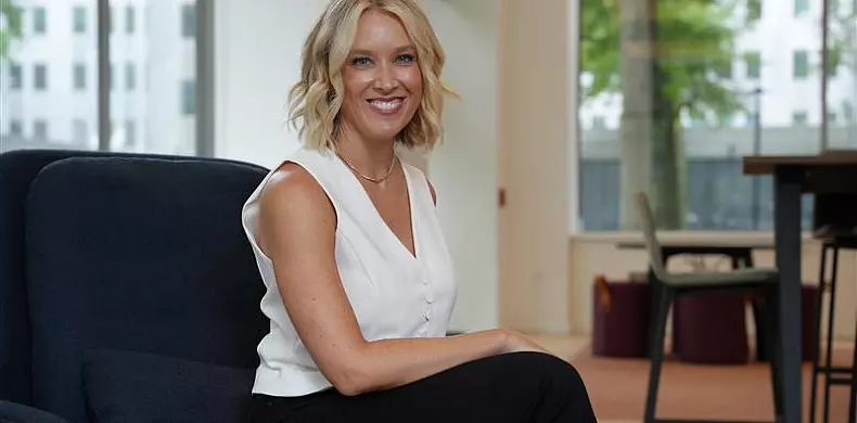 A woman with blonde hair in a white sleeveless top and black pants sits on a dark chair in a modern, bright office with large windows in the background. She is smiling at the camera.