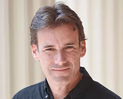 A man with short brown hair wearing a black collared shirt smiles slightly at the camera. The background is softly blurred with vertical beige and white stripes.