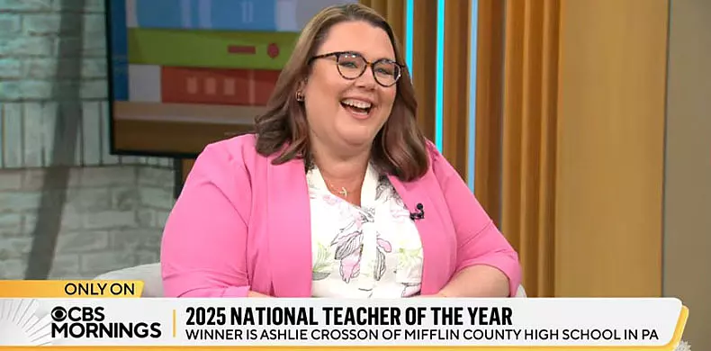 A woman wearing glasses and a pink blazer smiles while seated on a TV set. The screen text reads: "2025 National Teacher of the Year: Winner is Ashlie Crosson of Mifflin County High School in PA.
