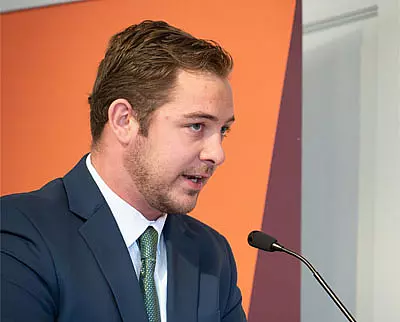 A man in a blue suit and green tie speaks into a microphone at a podium, with an orange and maroon background behind him.