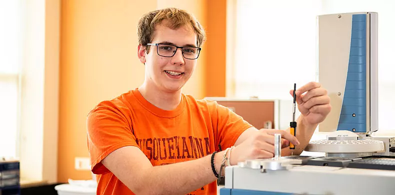 A young person wearing glasses and an orange "Susquehanna" T-shirt smiles while working with scientific equipment in a bright laboratory.