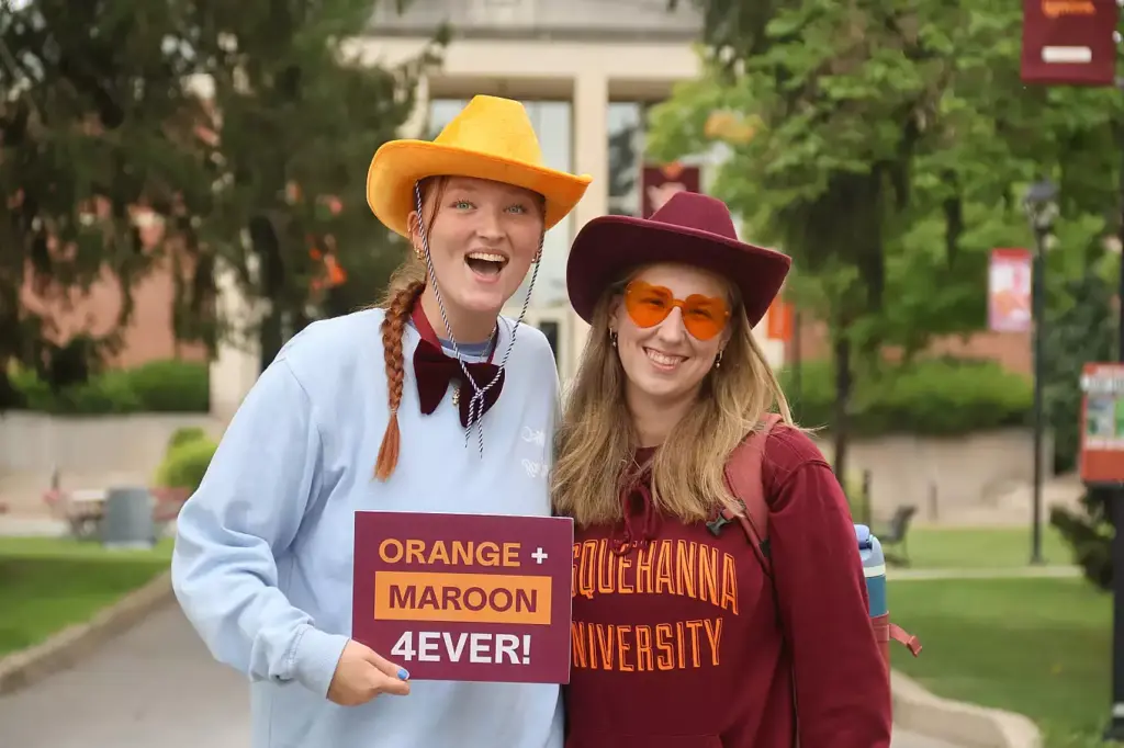 Two smiling people in cowboy hats and sunglasses pose outdoors, one holding a "ORANGE + MAROON 4EVER!" sign. Sporting a Susquehanna University sweatshirt, they celebrate campus pride and share enthusiasm for the many Ways to Give back.