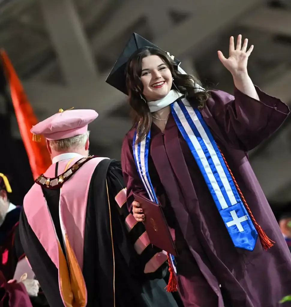 A female graduate in a maroon cap and gown smiles and waves while holding her diploma on stage. She wears a blue and white stole. An academic official in pink robes stands nearby, facing away.