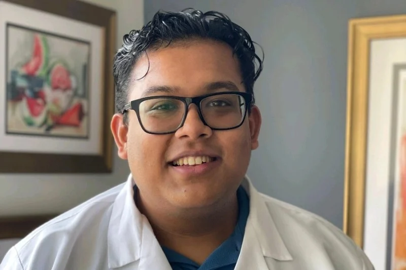 A person wearing glasses and a white medical lab coat stands indoors. The coat has logos from Harvard Medical School and Massachusetts General Hospital. Artworks are visible on the wall behind them.