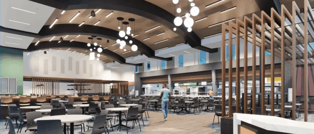 A modern cafeteria building with round tables and chairs, high ceilings adorned with decorative lighting, wooden accents, and large windows. The inviting space features a few people walking near the food counters in the background.