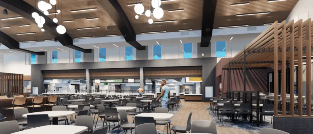 A spacious, modern cafeteria showcases the versatility of building and spaces, featuring high ceilings, round hanging lights, and many empty tables and chairs. A few people are standing near a food counter beneath bright windows.
