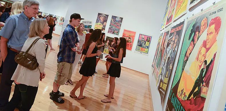 A group of people observe and discuss colorful vintage movie posters displayed on the walls of a brightly lit art gallery with a wooden floor.