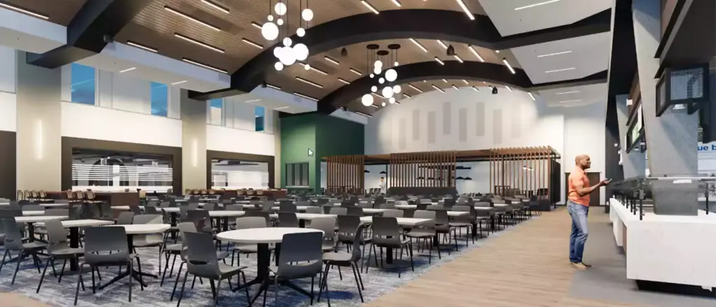 A spacious, modern cafeteria showcases impressive building and spaces design, with rows of round tables and chairs, high arched ceilings adorned with hanging lights, and one person standing at a counter on the right side of the image.