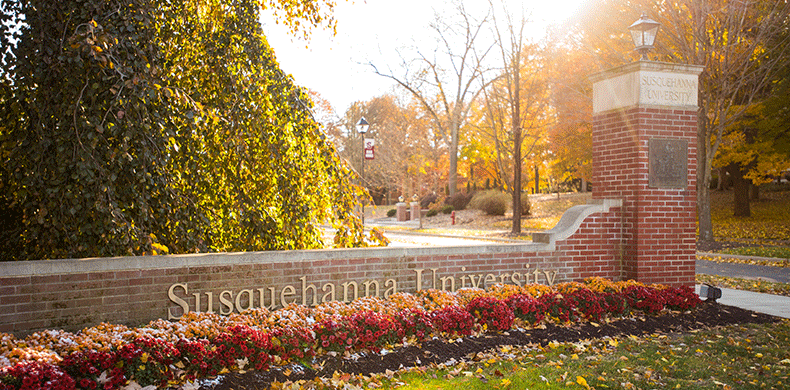 A brick entrance sign for Susquehanna University surrounded by colorful flowers and trees with autumn foliage, under bright sunlight.